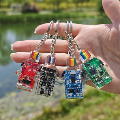 Four circuit board robot keychains in red, black, blue and green hanging from fingers outdoors