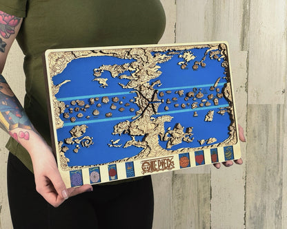 Person in a green top holds a light birch layered One Piece world map with blue sea and fruit panels against pale wood planks.