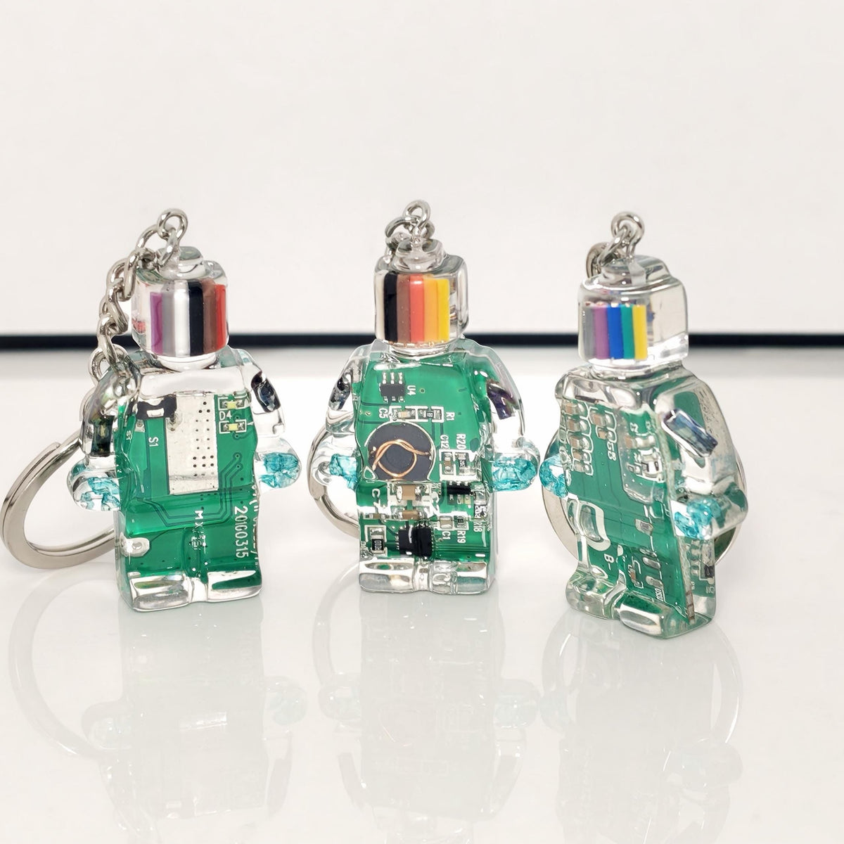 Three green circuit board robot keychains with striped heads and silver key rings on a white surface