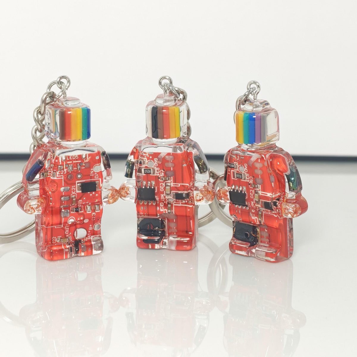 Three red circuit board robot keychains with multicoloured striped heads and silver key rings on a white surface