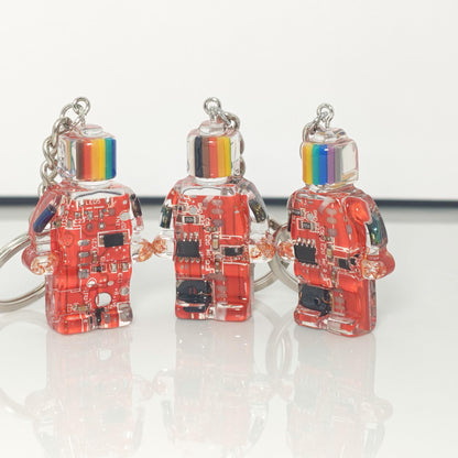 Three red circuit board robot keychains with multicoloured striped heads and silver key rings on a white surface