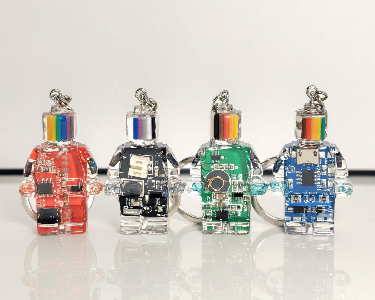 Four robot-shaped circuit board keychains in red, black, green and blue with striped heads on a white surface