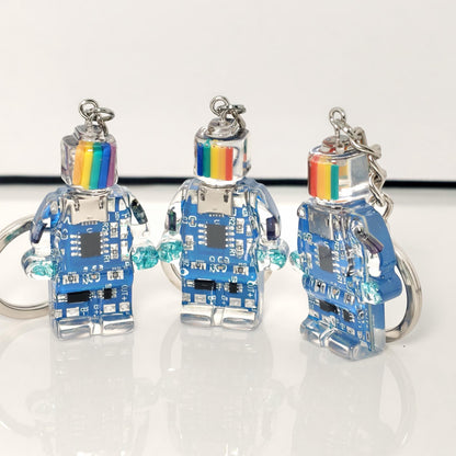 Three blue circuit board robot keychains with rainbow-striped heads and silver key rings on a white surface