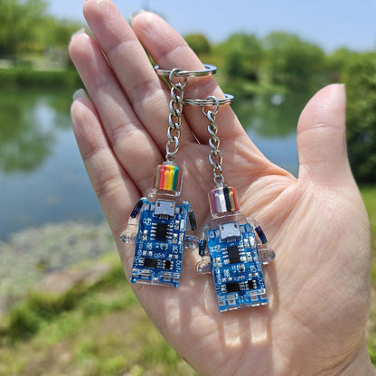 Two blue circuit board robot keychains on chains held in an open hand outdoors near a pond