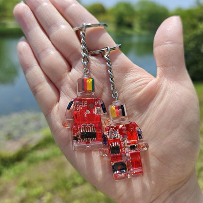Two red circuit board robot keychains on chains held in an open hand outdoors by a pond