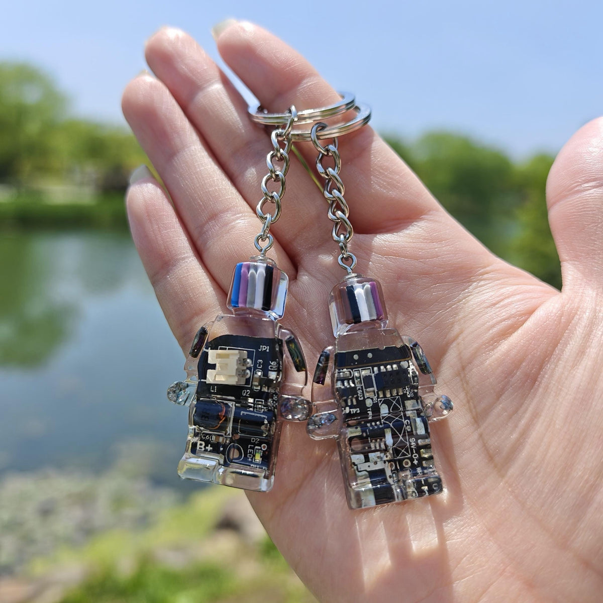 Two black circuit board robot keychains on chains held in an open hand outdoors by a pond