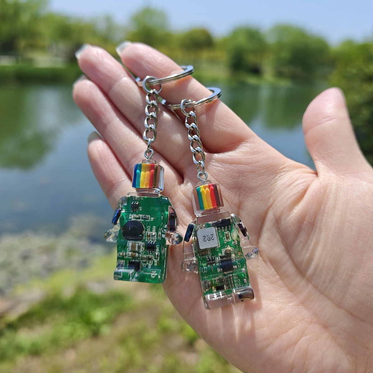 Two green circuit board robot keychains with rainbow-striped heads held in an open hand outdoors