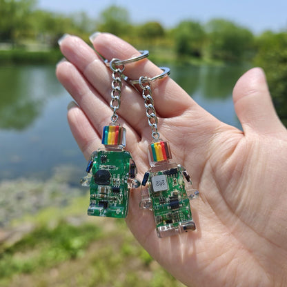 Two green circuit board robot keychains with rainbow-striped heads held in an open hand outdoors