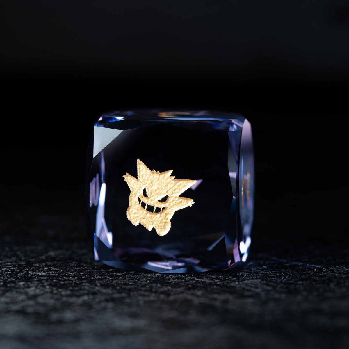 Faceted blue-violet clear d6 die with a gold grinning ghost-like creature symbol on its face, on a dark surface