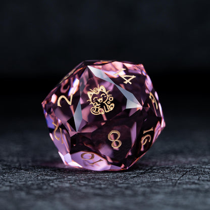 Faceted pink clear d20 die with gold numbers and a gold smiling cat-like creature symbol on a dark textured surface